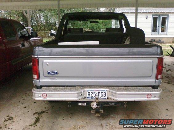 Silver and gray Ford Bronco rear view, spare tire in bed, parked next to a red truck