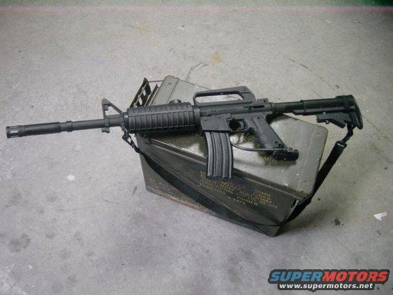 Black assault rifle with a carrying handle rests on a dark green ammo can. The can has some text on its side