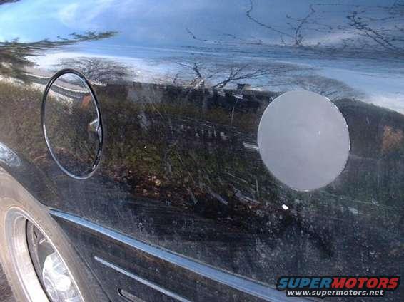 Black car body panel showing a fuel door and a circular dent. Reflections of trees and sky are visible