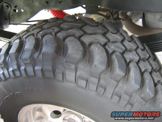 Close-up of a large off-road tire with aggressive tread, showing suspension components and exhaust pipe above