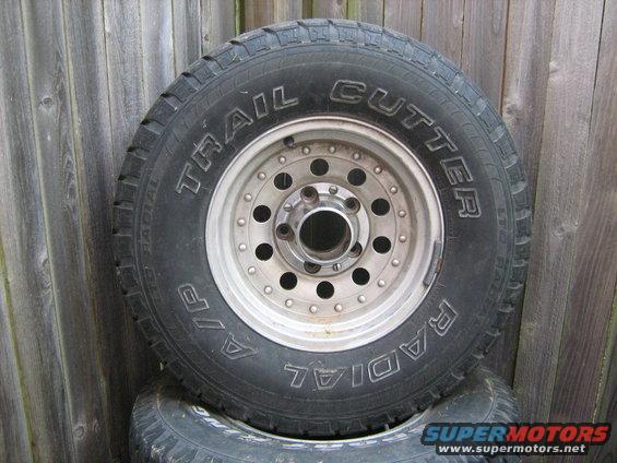 Tire with 'Trail Cutter' text and a silver wheel with multiple lug nut holes, leaning against a wooden fence