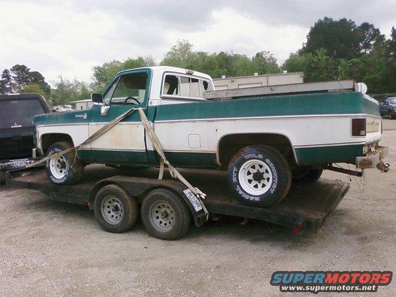 Green and white pickup truck on a trailer, strapped down. Tires have white lettering