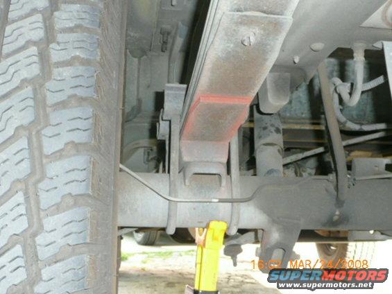 Close-up of a vehicle's rear axle and leaf spring suspension, with a tire visible on the left. A yellow jack is supporting the axle