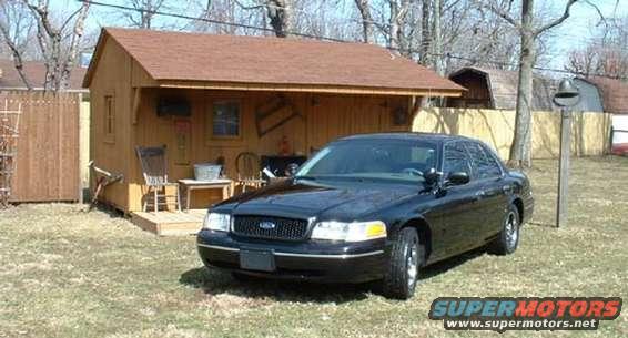Black sedan parked in front of a wooden shed. Watermark says SUPERMOTORS