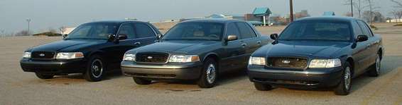 Three Ford Crown Victorias parked in a lot. The one in front is dark gray, the middle one is dark gray, and the one on the left is black