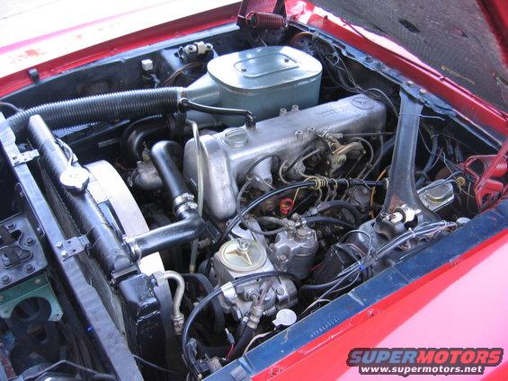 Engine bay of a red car, showing the engine, radiator, and air filter assembly. A Mercedes-Benz star emblem is visible on the engine