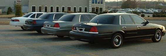 Four sedans parked in a lot, viewed from the rear. The closest is a black 1998 Ford Crown Victoria