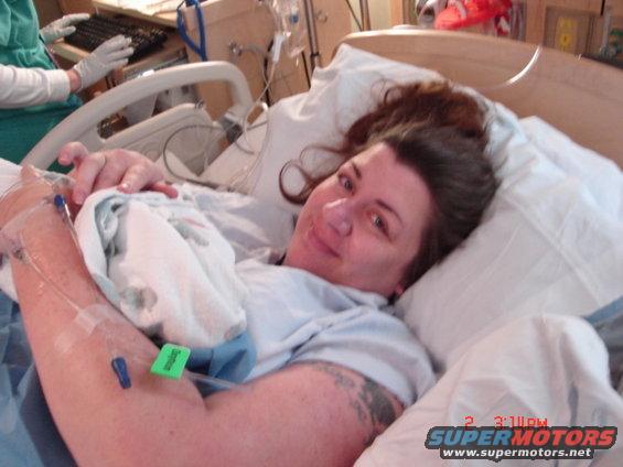 Woman in hospital bed with IV drip, looking at camera. Medical equipment and a person in scrubs are visible in the background