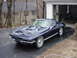 1964 Chevrolet Corvette - A dark blue coupe with distinctive hood scoops sits in a driveway, partially shaded by trees.