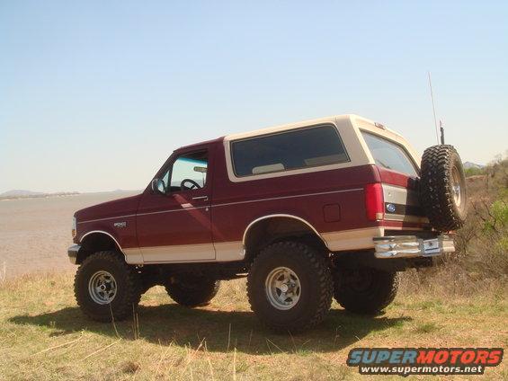Maroon and tan Ford Bronco parked on grassy hill near water. Spare tire mounted on rear