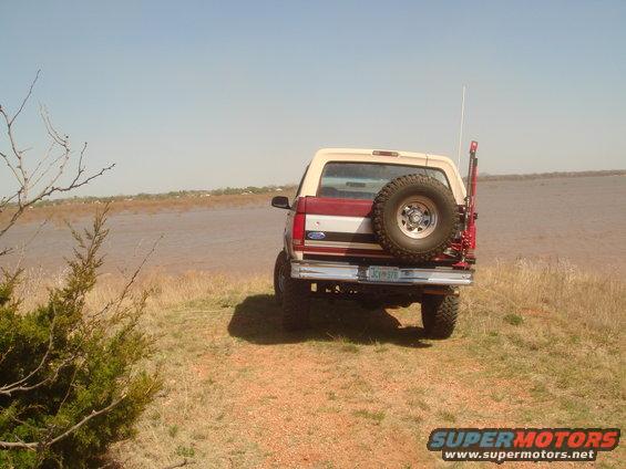 1992 Ford Bronco, rear view, red and tan, with spare tire mounted on back, parked on grassy bank near water
