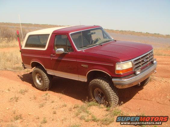 Red 1992 Ford Bronco with a white top, lifted suspension, and off-road tires, driving on a dirt path