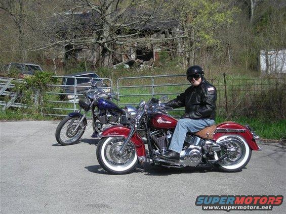 Man in leather jacket on red motorcycle, another motorcycle behind him. Old building in background