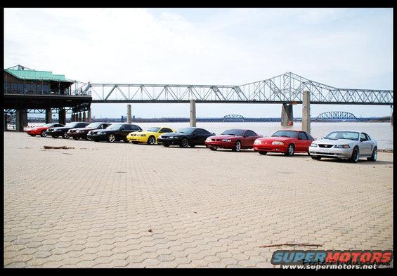 Several Ford Mustangs parked in a line by a river with a bridge in the background