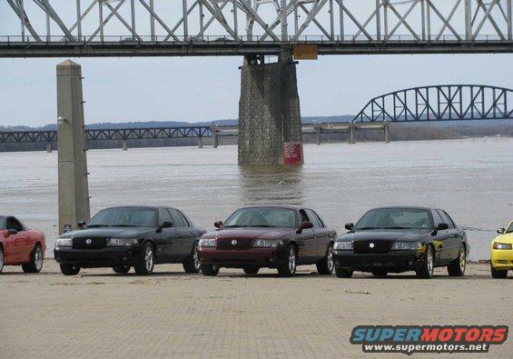 Three black and maroon 2003 Mercury Marauders and a red car parked on a sandy area near a bridge and river