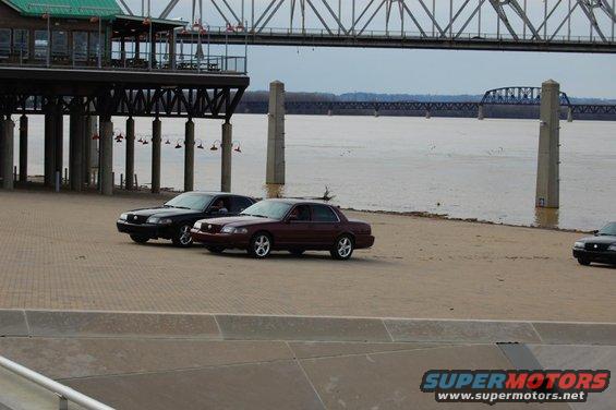 Two 2003 Mercury Marauders parked on a paved area near a river with a bridge in the background