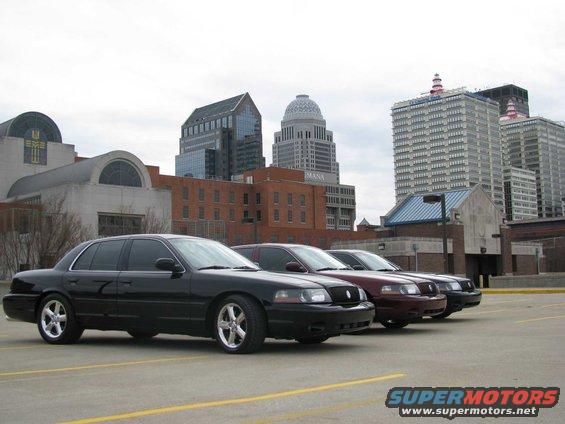 Black 2003 Mercury Marauder in foreground, followed by two other sedans, parked in a lot with city buildings