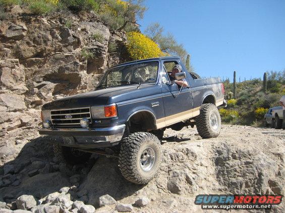 Blue Ford Bronco with tan stripe climbing rocks, driver visible