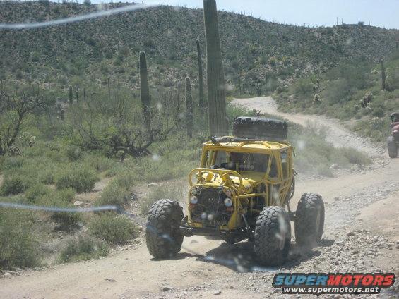 Yellow off-road buggy with a spare tire on top, driving on a dirt road in a desert landscape