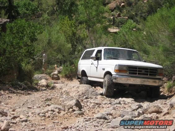 White Ford Bronco navigating a rough, rocky off-road path with sparse vegetation