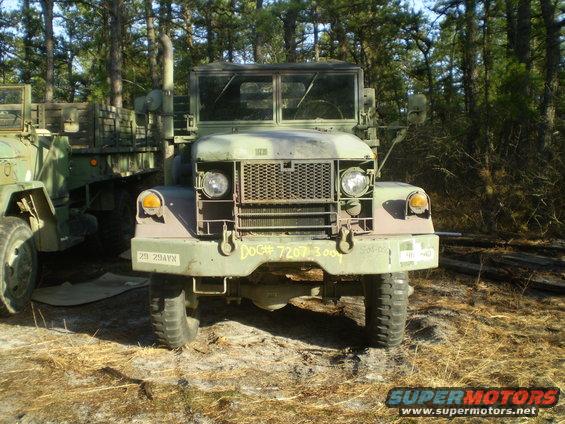 Front view of a camouflage military truck parked next to another military truck in a wooded area