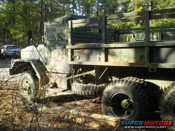 Military truck with wooden bed, parked outdoors. Another vehicle is visible in the background