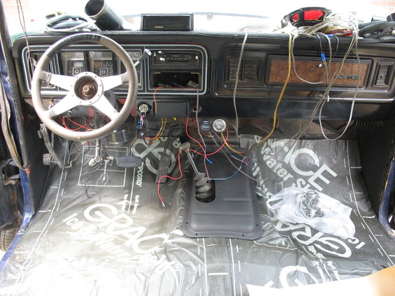 Interior of a vehicle with a steering wheel, dashboard, and center console. Lots of wires are visible, and the floor is covered with sound-deadening material