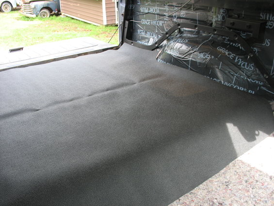 Gray carpeted interior of a vehicle cargo area with black sound dampening material on the walls. Another vehicle is visible outside