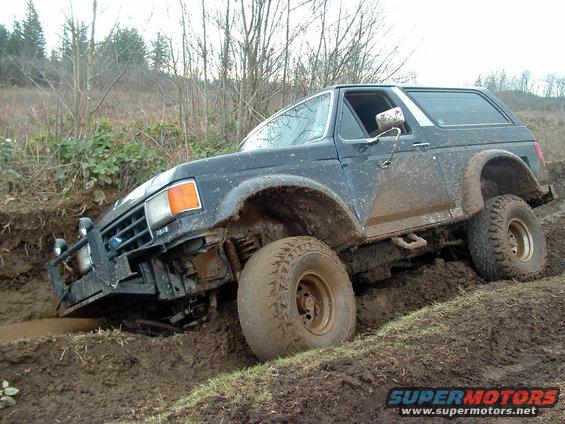 Mud-covered dark blue Ford Bronco with off-road tires stuck in mud, angled from the front-left