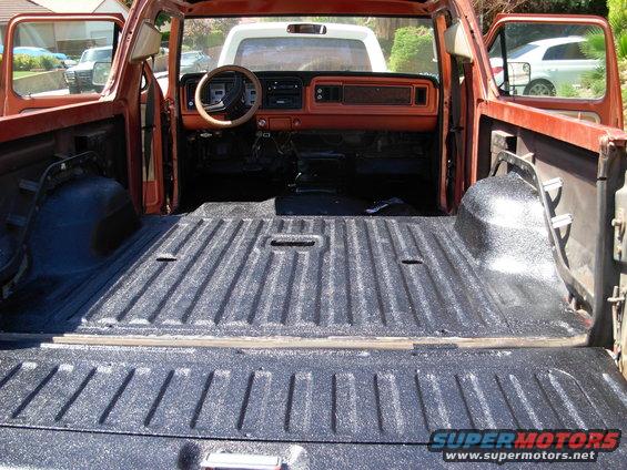 Interior and bed of an orange pickup truck, showing dashboard, steering wheel, and bed liner. Another white vehicle is visible in the background