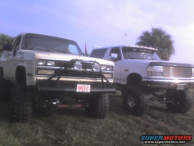 Two lifted trucks, one white Ford Bronco and a beige truck, parked on grass under a blue sky