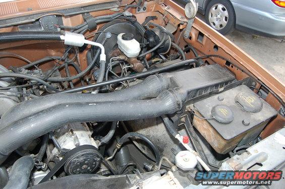 Engine bay with hoses, brake fluid reservoir, and air intake. Another vehicle is visible in the background