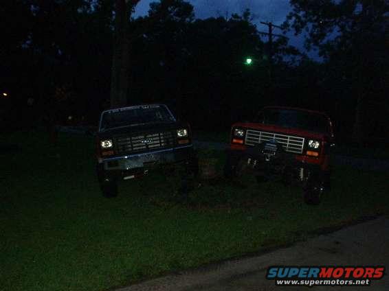 Two trucks parked on grass at night. The truck on the left is dark colored, the truck on the right is red