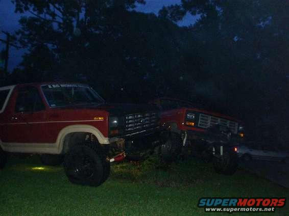 Two red SUVs parked on grass at dusk. The one in front has a black hood