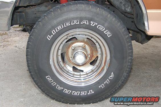 Close-up of a Liberator All Terrain tire on a rusty rim, showing part of a vehicle's frame and fender