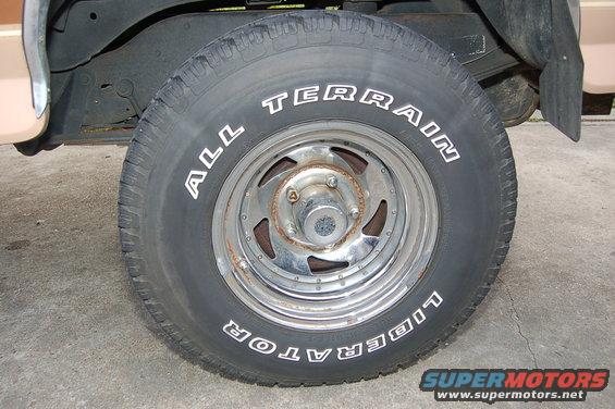 Tire with white lettering 'ALL TERRAIN' and 'LIBERATOR' on a chrome wheel. Part of a tan vehicle and black fender are visible