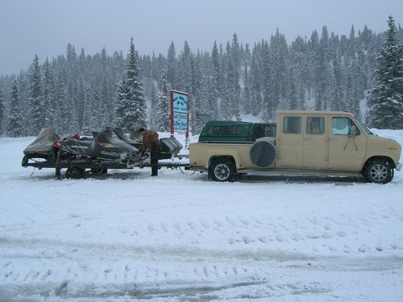 van towing snowmobiles on trailer in snow, trees in background