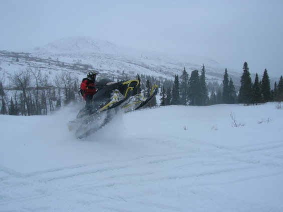 Snowmobile kicking up snow on a snowy hillside with trees and mountains in the background