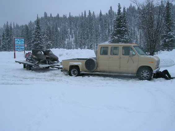 tan van with four doors towing a trailer with two snowmobiles, person working under the van in snow