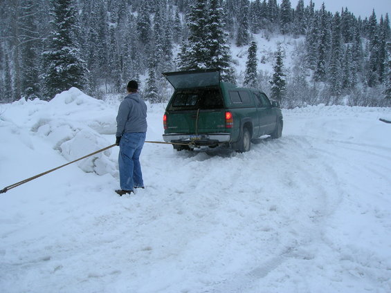 Green pickup truck with cap, rear view, in snowy area. Person holding a long pole near truck