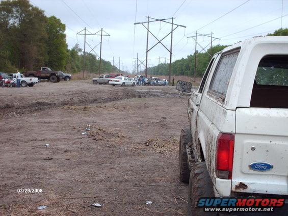 White Ford Bronco rear view, muddy terrain, power lines overhead. Multiple vehicles in background