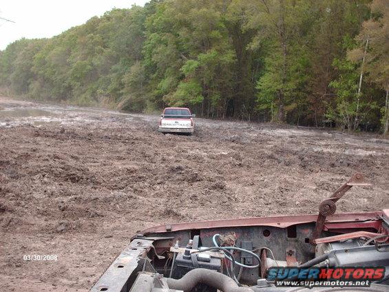 Muddy field with a red Chevrolet pickup truck stuck in the distance. Foreground shows the engine bay of another vehicle