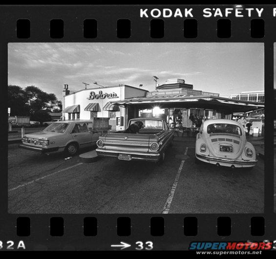 Ford Falcon Ranchero pickup truck with a dog in the bed, parked next to a VW Beetle outside a diner