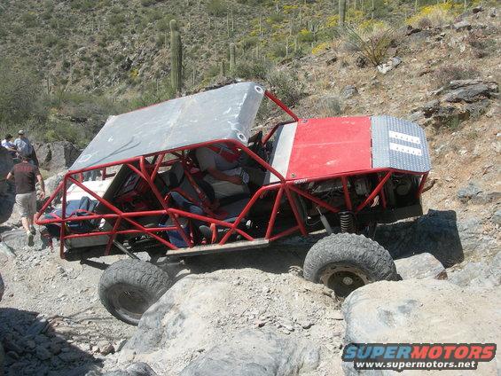 Red off-road buggy with a roll cage and metal roof, navigating a rocky desert terrain
