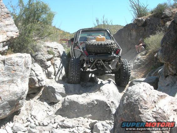Off-road vehicle with spare tire and jerry can on rear, climbing over large rocks on a narrow trail