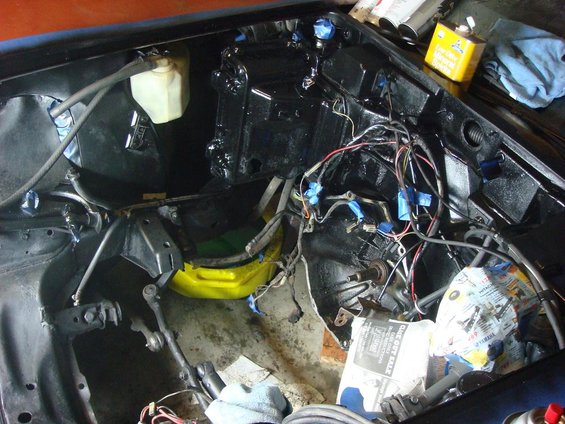 Car engine bay with wiring harness exposed, parts removed, and some components painted black. A yellow fluid container and rags are visible