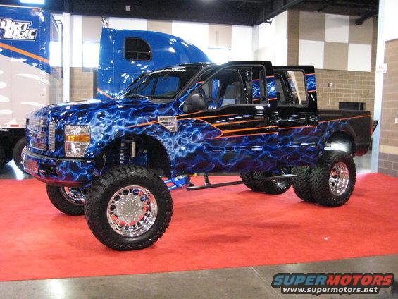 Lifted pickup truck with blue lightning graphics and orange stripes, parked indoors on a red carpet next to a semi-truck
