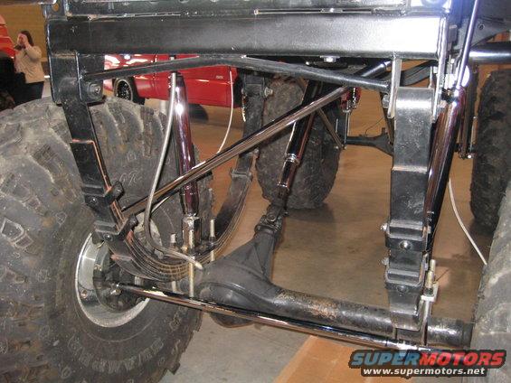 Close-up of the rear suspension and massive tires of a lifted off-road vehicle. Another red vehicle is visible in the background