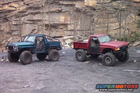 Green off-road truck with roll cage and red pickup truck with off-road tires parked on dirt near rocky cliff