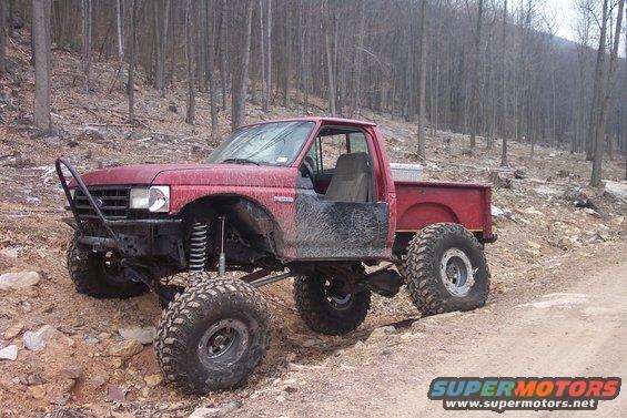 Red lifted 1987 Ford F-150 pickup truck with off-road tires and no doors on a dirt trail